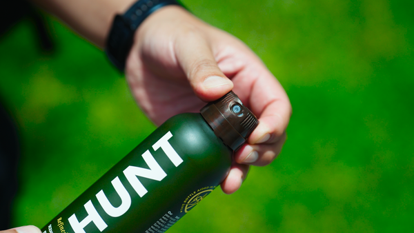 Hand holding a Hunt Refined Repellent Spray bottle against a background of green grass.