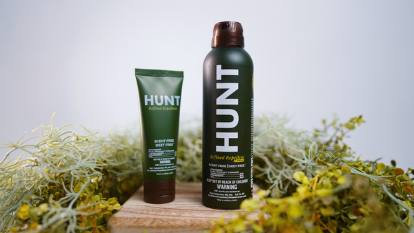 HUNT Refined Repellent Lotion and Spray on a wooden block with foliage in the background.