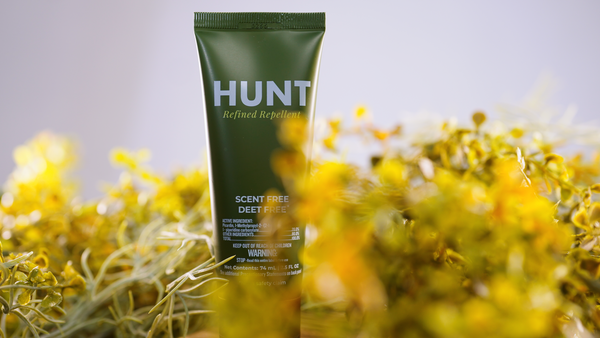 Tube of Hunt Refined Repellent Lotion surrounded by yellow flowers on a light gray background.