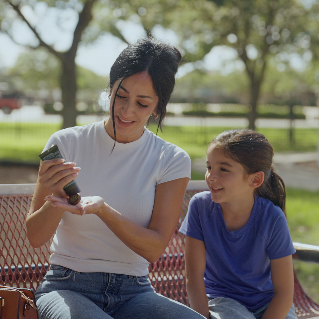 A photo of a mom and daughter in a park. The mom is applying HUNT Refined Repellent to her daughter.